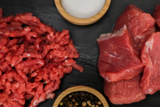 A close-up view from above of chunks of beef meat and red minced meat with streaks of fat lying on a black background with spices and salt in wooden bowls on the sides. The concept of cooking, shredding meat, diet, natural products. High quality photo