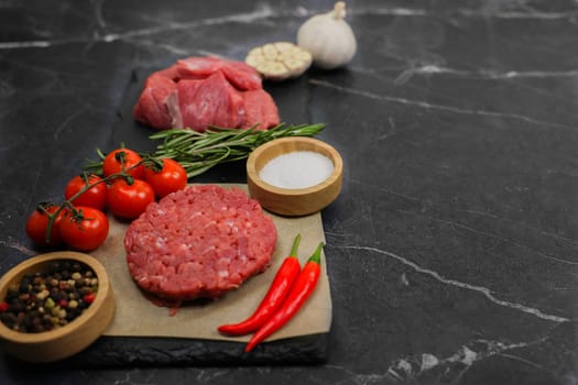 Close-up on the left is a raw beef round patty lying on paper surrounded by seasonings and vegetables, behind them separated by a sprig of rosemary lies meat cut into chunks on a dark background with space for text. High quality photo