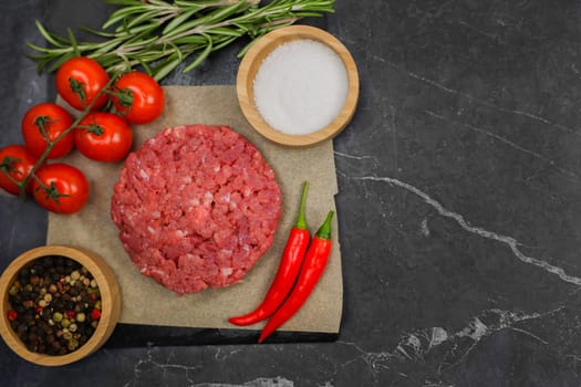 Close-up left of raw beef patty lying on paper surrounded by salt, pepper, seasoning, red hot pepper and tomatoes on a branch on a dark background with space for text. High quality photo
