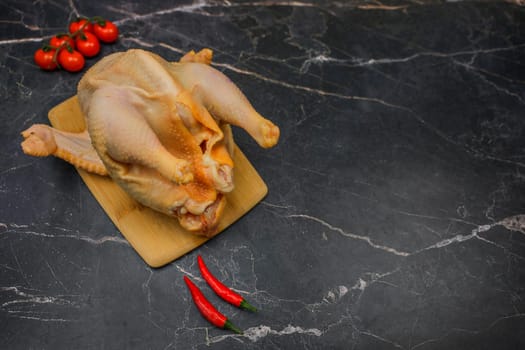A chicken carcass, butchered and plucked, lies on a wooden board against a background of black marble, surrounded by cherry tomatoes and hot red peppers with space for text. The concept of cooking, poultry meat, dietary dishes. High quality photo