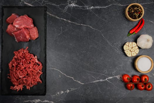 Top view of chunks of beef and minced meat lying on a black cutting board on the left, vegetables and condiments in wooden bowls on a dark background with space for text on the right. The concept of cooking, shredding meat, diet, natural products. High quality photo