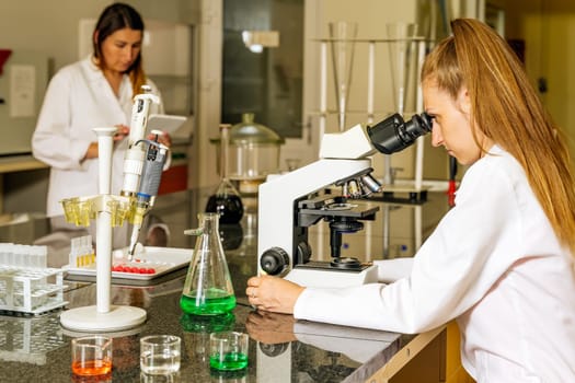 Two female laboratory technicians researching covid 19 samples under microscope and uploading data through tablet for coronavirus vaccination