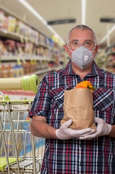 Coronavirus protective measures. Man with protective mask, safety goggles and medical gloves carries a bag of fresh vegetables over blurred grocery store. Avoiding social contact and hold distance.