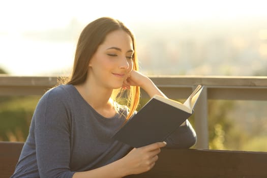 Relaxed woman reading a hard cover book at sunset sitting on a park bench