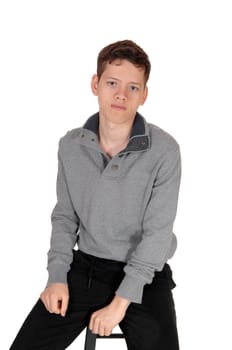 A happy young teenage boy sitting on a chair in the studio in a gray.sweater, looking into the camera, isolated for white background.