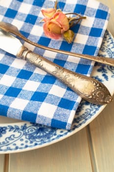 Silver fork and knife with an empty vintage plate on a blue napkin, with dry pink rose