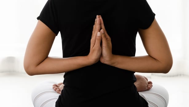 Asian women doing yoga meditating exercise at home, sitting in Lotus pose or Padmasana with raised hands namaste behind the back in white bedroom. Working out wearing black shirt and white pants. Healthcare concept.