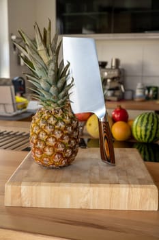 A cutting board with a whole pineapple fruit and a cleaver standing on a worktop in the kitchen in the background are more fruits and kitchen utensils