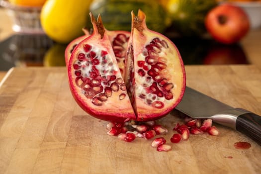 Close up of two half pomegranates fruits and a damascus kitchen knife on a wooden cutting board with pomegranate seeds in the foreground in a domestic kitchen
