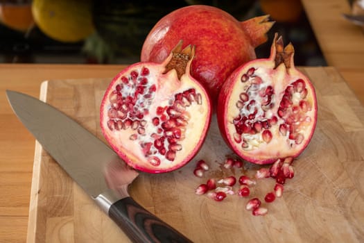 Close up of one whole and two half pomegranates fruits and a damascus kitchen knife on a wooden cutting board with pomegranate seeds in the foreground in a domestic kitchen