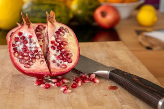 Close up of two half pomegranates fruits and a damascus kitchen knife on a wooden cutting board with pomegranate seeds in the foreground in a domestic kitchen