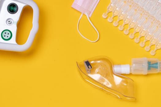 Coronavirus protective measures. Closeup of a inhaler device with medical face mask or nebulizer for treatment of the respiratory tract, sodium chloride in ampoules and a surgical mask flat lay on yellow background. Prevention for COVID-19 infection by inhaling saline.