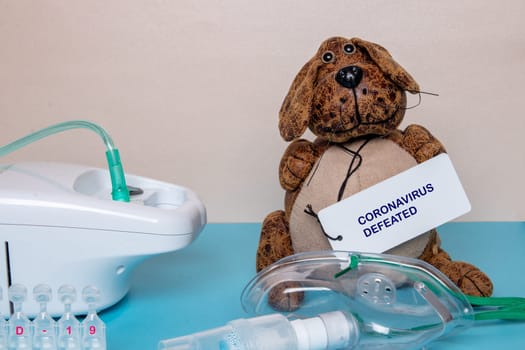 Funny dog with a coronavirus defeated tag a medical face mask or nebulizer for treatment of the respiratory tract, further ampoules and a inhaler device on a blue table over bright background.