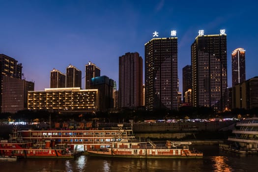 Chongqing, China -  August 2019 : Night view of the Chongqing city lights over Jialing and Yangtze river with luxury cruise passenger boat docked on the shore and illuminated skyscrapers in the background