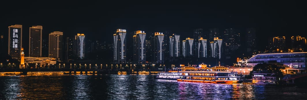 Chongqing, China -  August 2019 : Night view of the Chongqing city lights over Jialing and Yangtze river with luxury cruise passenger boat docked on the shore and illuminated skyscrapers in the background