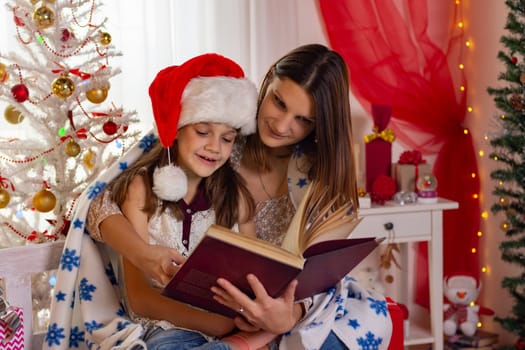 Mom and daughter sit next to a Christmas tree and read a big book