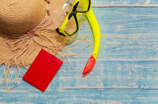 Flat lay summer traveler accessories of straw beach hat, yellow snorkel and red passport on blue wooden background. Summer vacation and travel concept.