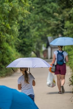 A vertical shot of a child following her mother on a beautiful tree-covered street with an umbrella