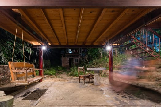 A beautiful picture of a person swinging back and forth on a swing in a hut surrounded by foliage trees at night