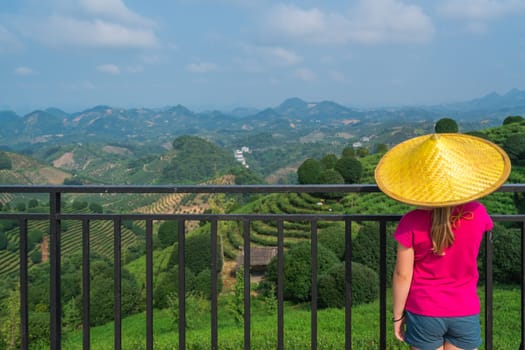 A beautiful closeup of a female leaning on the fences in Asian conical hat against a foliage forest and mountains