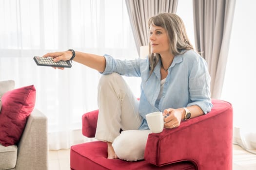Mature woman in armchair with TV remote and coffee