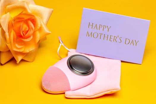 Mothers day greeting card. Close-up of a modern electric face cleansing brush a beautiful yellow blossom of flower and a tag with inscription Happy mothers day on yellow background. Beauty for the mom. Macro.