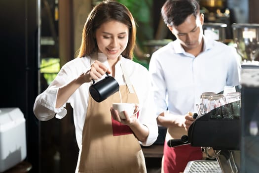 Young adult asian female woman barista pouring fresh milk to prepare latte coffee for customer in cafe bar with her colleague working in background. For small business startup in food industry concept
