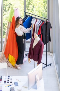 Top view of young adult asian fashion designer working from home and standing near clothing rack or clothes horse with colorful and fashionable shirts. Using for entrepreneur startup concept.