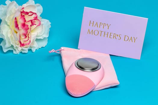 Mothers day greeting card. Close-up of a modern electric face cleansing brush a beautiful white blossom of flower and a tag with inscription Happy mothers day on blue background. Beauty for the mom. Macro.
