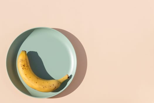 Minimalist image of a banana on a light green plate with a beige background and hard shadows of daylight. Top view.