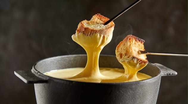 Delicious melted cheese fondue in winter served in a cast iron pot with forks dipping toasted baguette and rising steam over a dark background
