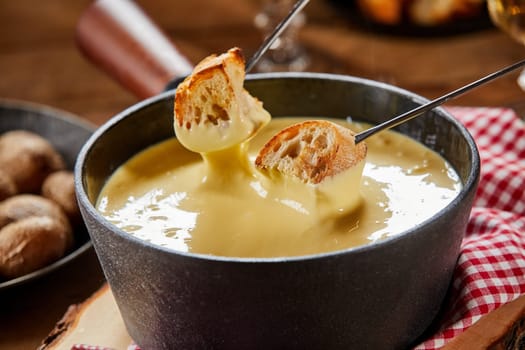 Two forks with toasted baguette being dipped into a pot of traditional Swiss cheese fondue with raclette in close up