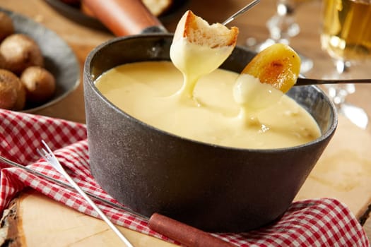 Steaming pot of creamy cheese fondue with grilled baby potatoes and toasted baguette for dipping in a close up tilted view on a rustic table