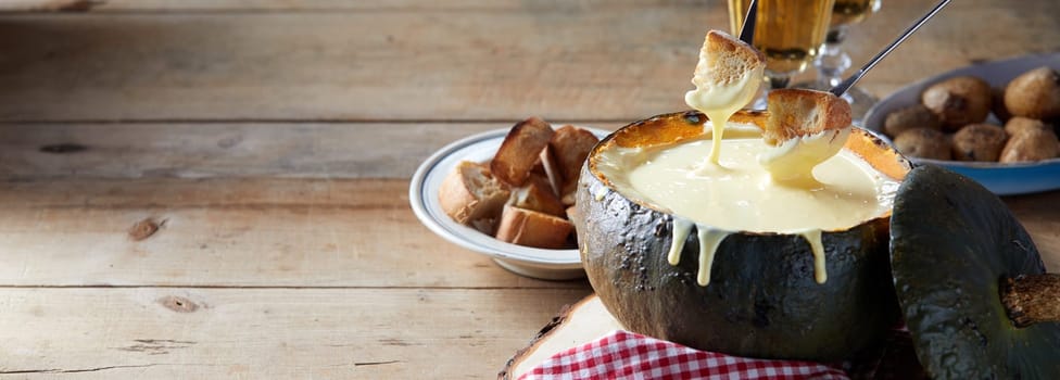 Panorama banner with a delicious traditional Swiss cheese fondue served in a hollwed out pumpkin on a rustic table with accompanying toasted bread and potatoes, with copy space