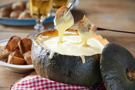 Toasted baguette dipped into melted cheese fondue served in a hollow autumn pumpkin on a rustic table in close up