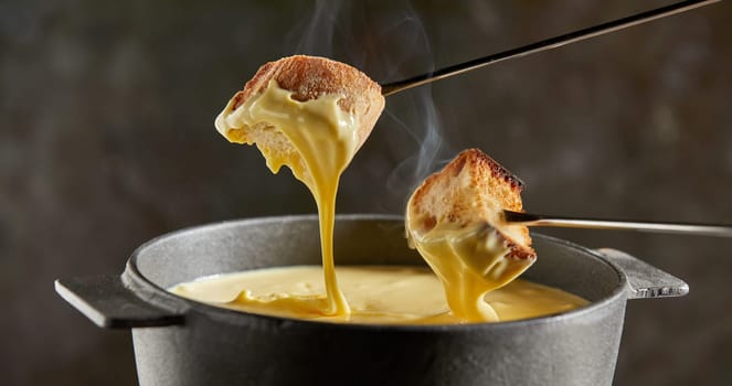 Dipping toasted bread into a hot cheese fondue dripping from the forks in a close up view on a dark background with steam