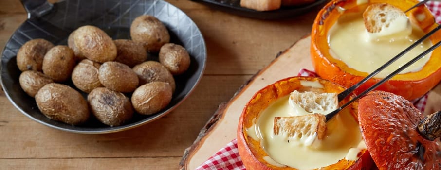 Servings of cheese fondue in pumpkin gourds viewed high angle on a rustic table with toasted bread and potatoes in a panorama banner