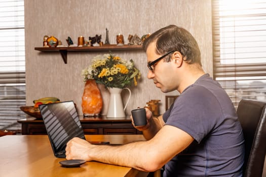 Middle age man drinking espresso and working on laptop from comfort of his home at sunny day during covid 19 or coronavirus pandemics and quarantine