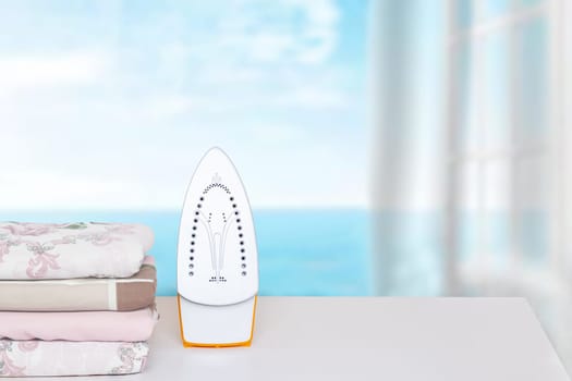 Household laundry ironing. Close-up of a yellow electrical iron and a stack of ironed clothes on an ironing board indoors over abstract blurred natural ocean background. Advertising for the laundry service with space.