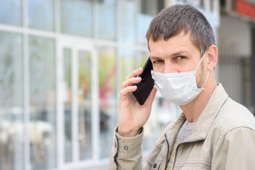 Portrait of a man in a medical mask with a mobile phone on the background of the store, looking in frame