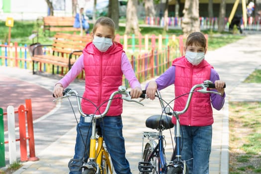 Two girls with medical masks on their face walk with bicycles in the playground