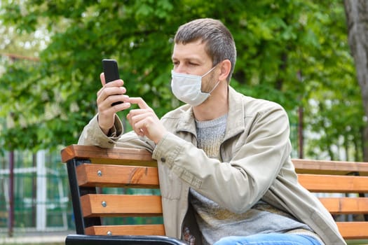 A man in a medical mask with a smartphone sits on a bench in the park