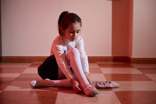 A young ballerina in a studio ties her ballet shoes, surrounded by warm, focused lighting, symbolizing dedication and the beauty of dance.