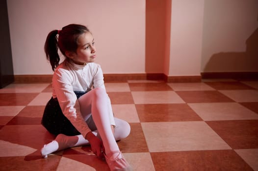 A girl wearing a ballet outfit sits on a tiled floor illuminated by warm light, capturing a moment of calmness and reflection.
