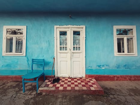 Walking stick, and a pair of old shoes and a chair on the doorstep of an aged house. Traditional rural building facade. Blue lime painted walls, white colored door and windows. Vintage home details.