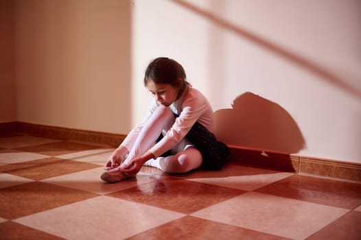 A young girl practices ballet, sitting gracefully on a checkered floor, illuminated by soft sunlight streaming into the room.