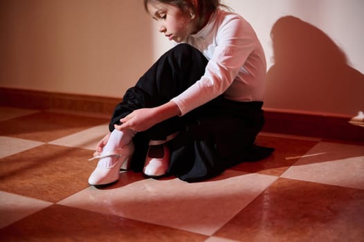 A young girl in elegant attire focuses on fastening her white shoes. The warm sunlight creates a relaxing atmosphere.