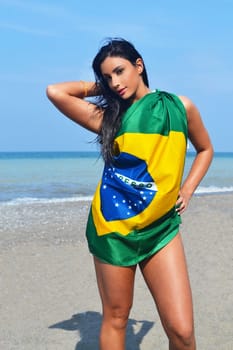 Beautiful young woman standing on the beach covered in a Brazilian flag.with her long black hair.