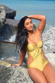 A beautiful young woman sunbathing on the beach sitting in a swimsuit.on some big rocks in a beige swimsuit, eye closed, sunbathing.