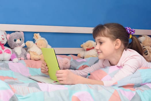 The girl lies in bed at home on her stomach and looks at the screen of the tablet